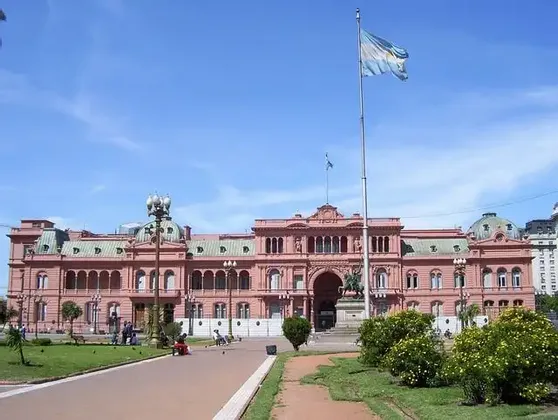 Un grande edificio governativo rosa si staglia dietro un parco, con una bandiera argentina che sventola su un'alta asta sotto un cielo azzurro intenso.