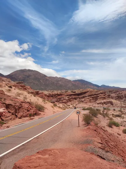 Una strada asfaltata con un cartello di limite di velocità si snoda attraverso un paesaggio desertico di formazioni rocciose rosse e montagne sotto un cielo blu e nuvoloso.