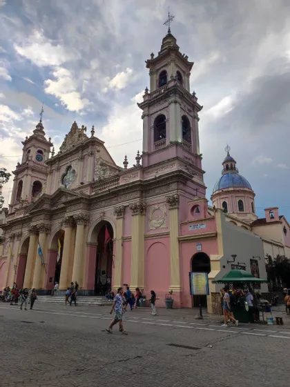 Una cattedrale rosa riccamente decorata, con colonne color crema e due campanili gemelli, si erge su una piazza acciottolata mentre le persone passeggiano.