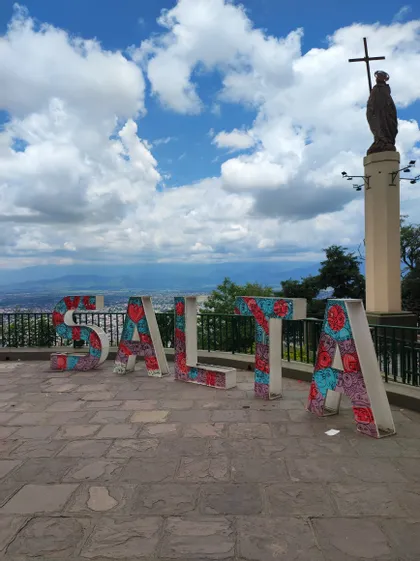 Un'insegna colorata che forma la parola SALTA su un punto panoramico in pietra, con una statua su un pilastro che domina un paesaggio urbano sotto un cielo nuvoloso.