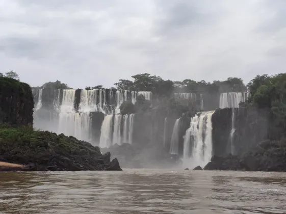 Una cascata ampia e a più livelli scorre su scogliere rocciose sormontate da alberi, riversandosi in un fiume sottostante sotto un cielo nuvoloso.