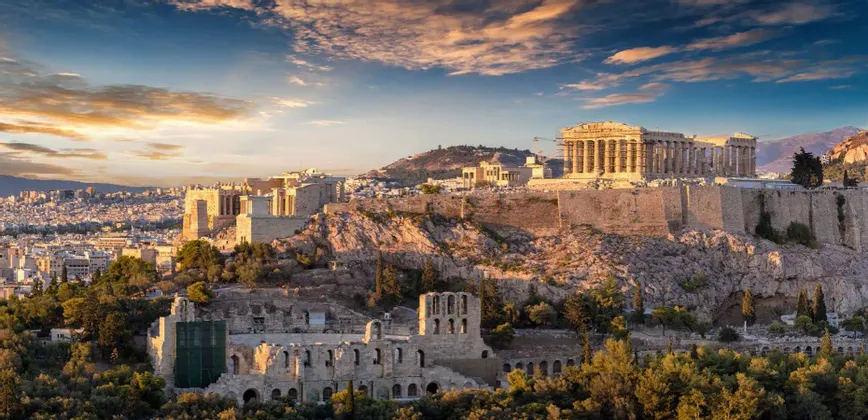 Eine historische Akropolis mit antiken Ruinen thront auf einem felsigen Hügel und blickt auf eine weitläufige Stadt unter einem farbenprächtigen Sonnenuntergangshimmel.