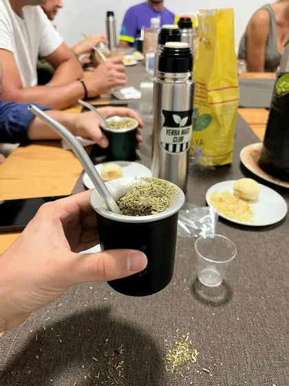Primo piano di una mano che sorregge una tazza tradizionale di yerba mate, mentre un gruppo WeRoad condivide la bevanda attorno a un tavolo.