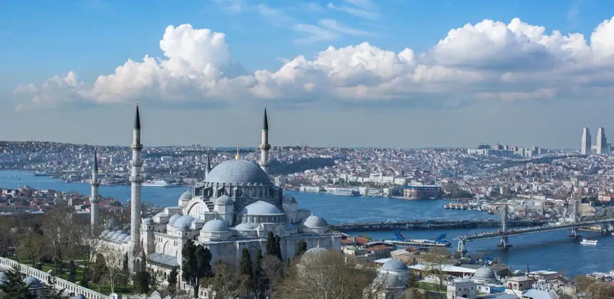Une grande mosquée avec plusieurs dômes gris et minarets surplombe un horizon urbain animé à travers une large étendue d'eau.