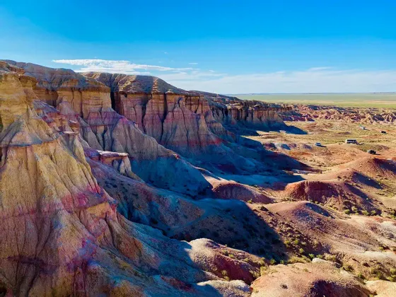 Formazioni rocciose colorate e stratificate e calanchi in un vasto paesaggio sotto un cielo azzurro limpido, con veicoli in lontananza.