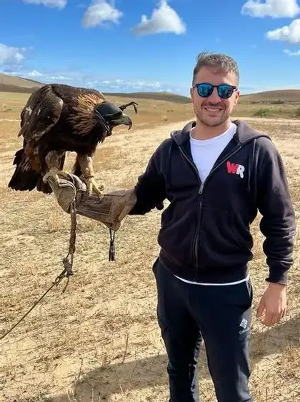 Un uomo con occhiali da sole sorride mentre tiene una grande aquila incappucciata su un guanto di cuoio in un paesaggio arido e collinare.