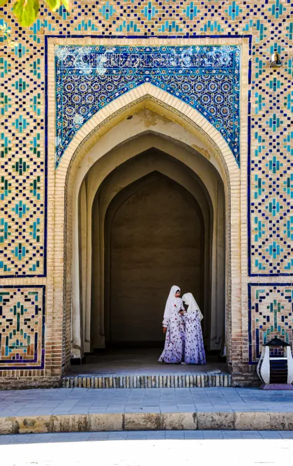 Zwei junge Mädchen in weißen Hijabs und passenden Kleidern stehen in einem kunstvollen Ziegelbogen, der mit blauen Mosaikfliesen verziert ist.