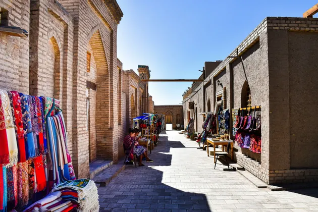 Eine sonnige Marktgasse mit bunten Textilien zum Verkauf entlang alter Backsteingebäude unter strahlend blauem Himmel.