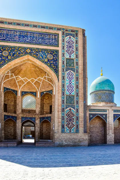 Die verzierte Ziegelfassade eines Gebäudes mit aufwendigen blauen, gelben und weißen Mosaikfliesen und einer türkisen Kuppel unter einem klaren blauen Himmel.