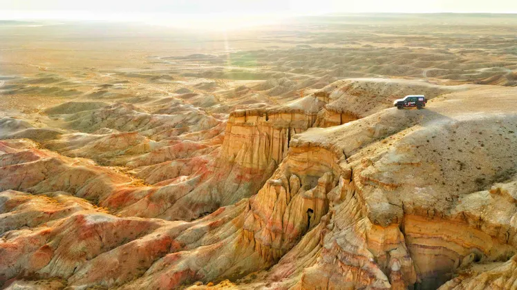 Vista aerea di un SUV parcheggiato su una scogliera che si affaccia su un paesaggio desertico di canyon erosi e colorati all'alba.