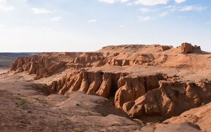 Una persona solitaria si trova sul bordo di un vasto canyon di roccia rossa sotto un cielo azzurro pallido con nuvole leggere.