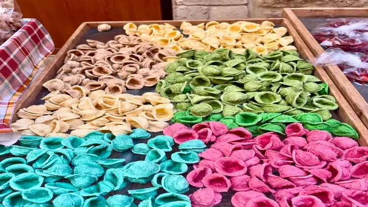 Pile colorate di orecchiette fresche, divise per colore ed esposte in vassoi di legno.