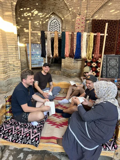 Un gruppo WeRoad in viaggio si siede con una donna locale su una panchina, apprendendo un mestiere in un laboratorio con filati colorati.