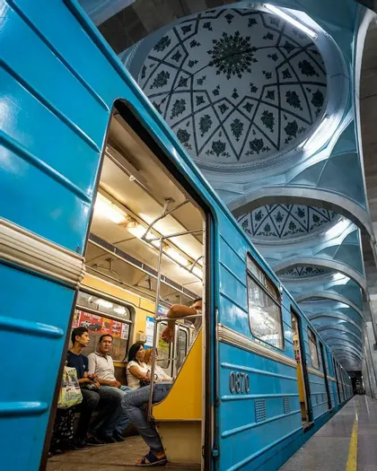 Un treno blu è fermo su una banchina della stazione, rivelando i passeggeri all'interno, sotto un soffitto a cupola ornato con motivi geometrici.