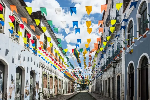 Bandiere colorate da festival e una lanterna di carta pendono sopra una stretta strada acciottolata tra vecchi edifici sotto un cielo parzialmente nuvoloso.
