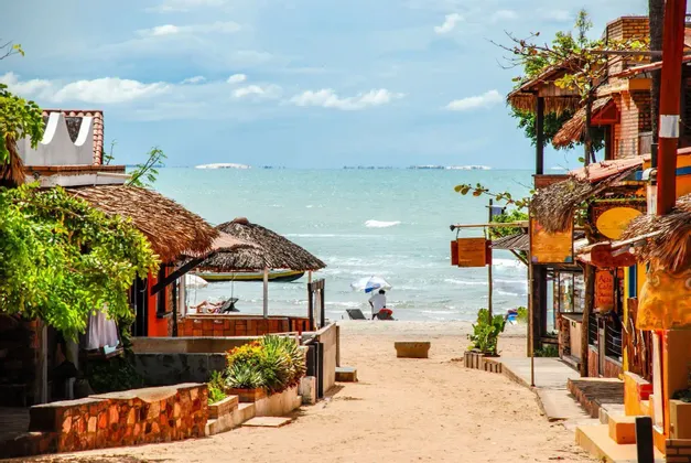Un sentiero sabbioso tra edifici rustici con tetti di paglia conduce a una spiaggia oceanica tranquilla sotto un cielo parzialmente nuvoloso.