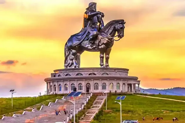 Una massiccia statua equestre metallica si erge in cima a un edificio circolare su una collina erbosa al tramonto, con persone sulle scale sottostanti.