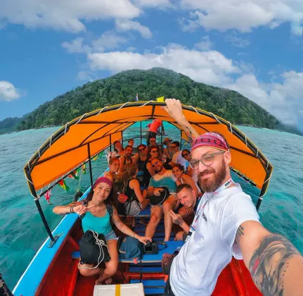 Un hombre se toma una selfie de su viaje en grupo de WeRoad en un barco de cola larga con un toldo naranja, con una exuberante isla tropical de fondo.