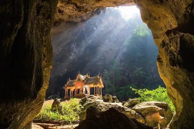 Un pabellón dorado ornamentado se alza dentro de una vasta cueva, iluminado por un rayo de sol desde una abertura superior.