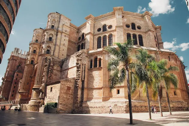 Una toma en contrapicado de una gran y ornamentada catedral de piedra con torres junto a una plaza pública con palmeras.