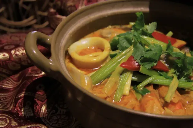 Un primo piano di un curry di mare con anelli di calamaro, sedano e coriandolo, servito in una ciotola di ceramica rustica su una tovaglia fantasia.