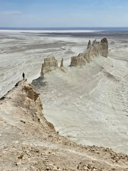 Una persona si trova su un'alta scogliera che domina un vasto paesaggio brullo con imponenti formazioni rocciose bianche.