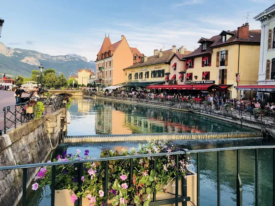 Un canal traverse une ville avec des bâtiments colorés et des restaurants avec terrasses, avec des montagnes visibles en arrière-plan.