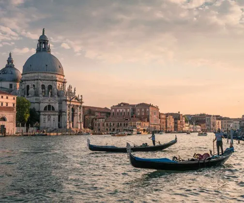 Gondole galleggiano su un canale di fronte a una grande basilica a cupola e a edifici storici al tramonto.