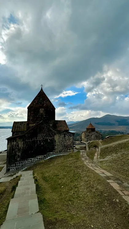 Due antiche chiese in pietra su una collina erbosa che dominano un lago, con montagne sullo sfondo sotto un cielo nuvoloso.