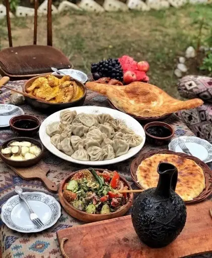 Un tavolo apparecchiato all'aperto con un banchetto di piatti tradizionali georgiani come i khinkali, pane, insalata, stufato e frutta fresca.