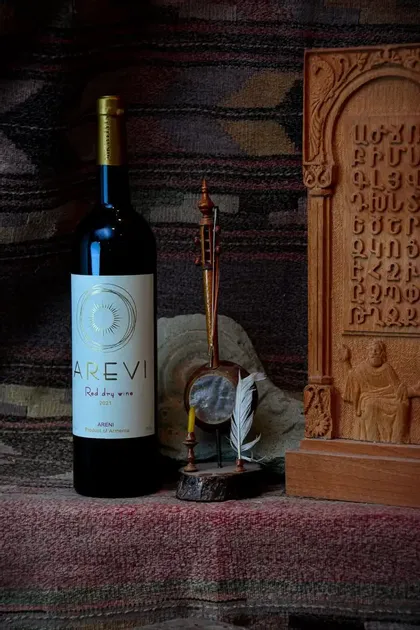 Una bottiglia di vino rosso armeno accanto a uno strumento a corda tradizionale e una tavoletta di legno intagliata su un tappeto tessuto.