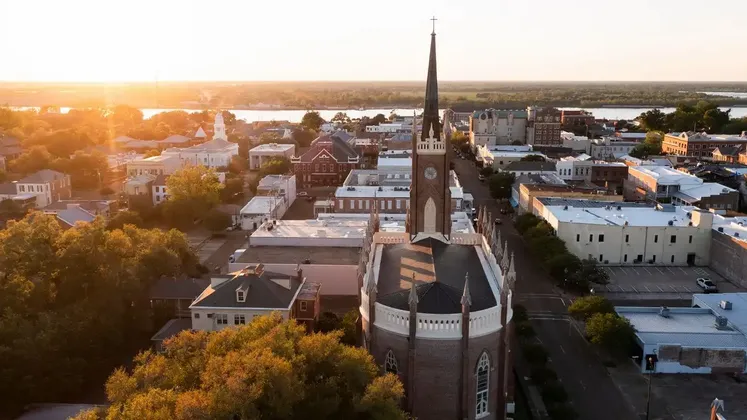 Una vista aerea di una città con una chiesa in mattoni e campanile al tramonto, con un ampio fiume sullo sfondo.