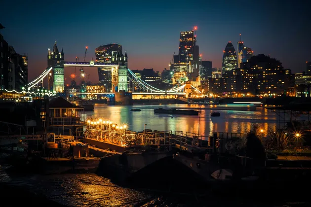 Un ponte basculante illuminato e lo skyline della città di notte, visti da oltre un fiume con barche ormeggiate in primo piano.