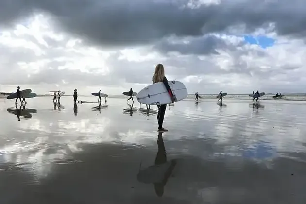 Eine WeRoad-Gruppe von Surfern mit Surfbrettern geht an einem nassen Strand entlang, ihre Spiegelungen sind im Sand sichtbar.