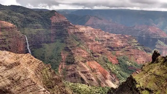 Ein dünner Wasserfall stürzt eine Klippe hinab in eine weite, zerklüftete Schlucht mit geschichteten roten Felsen und grünem Laub.