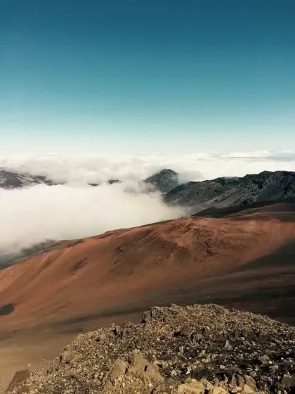 Ein Blick von einem felsigen Berggipfel hinunter auf einen rotbraunen Hang und ein Tal, gefüllt mit einem Wolkenmeer.
