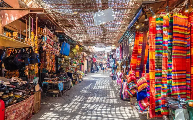 Un vicolo stretto del mercato con bancarelle che vendono tessuti colorati, borse e sandali sotto una tettoia intrecciata che proietta ombre disegnate.