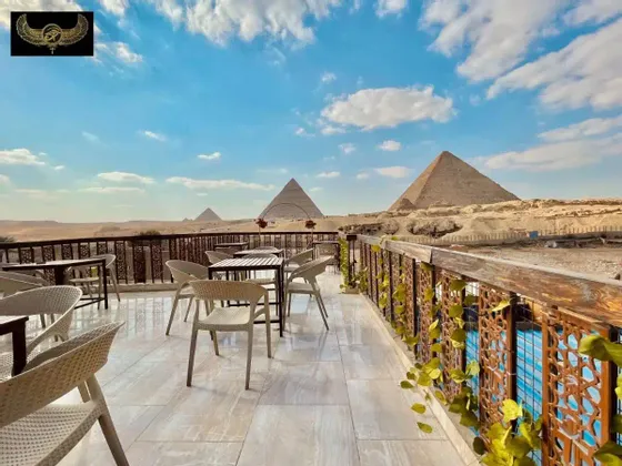 Una terrazza all'aperto con tavoli e sedie vuoti che si affaccia su tre grandi piramidi nel deserto sotto un cielo parzialmente nuvoloso.