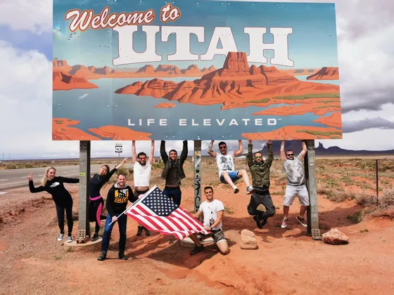 Membri di un viaggio di gruppo WeRoad si aggrappano e posano sotto un grande cartello 'Welcome to Utah', con una persona che tiene una bandiera americana.
