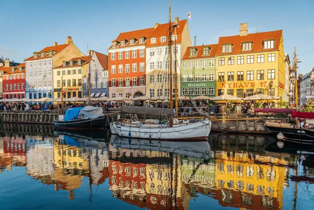 Edifici colorati e barche a vela ormeggiate si riflettono nell'acqua calma di un canale sotto un cielo azzurro chiaro.