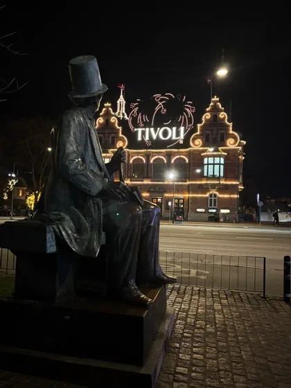 Una statua di bronzo di un uomo con cappello a cilindro è seduta su una panchina che si affaccia sul palazzo illuminato di Tivoli di notte.