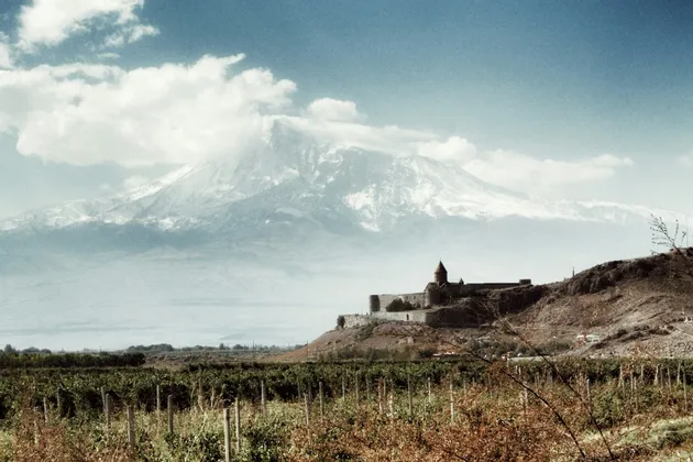 Un monastero in pietra si erge su una collina che domina un vasto vigneto, con una grande montagna innevata sullo sfondo sotto un cielo blu.