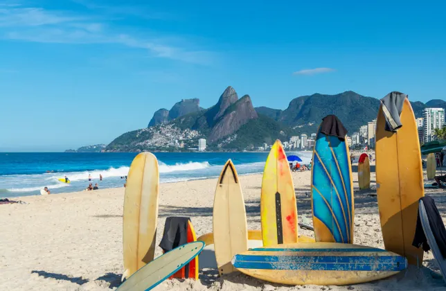 Une rangée de planches de surf colorées est plantée dans le sable sur une plage, avec l'océan, une ville et des montagnes en arrière-plan.
