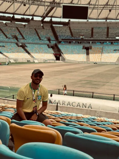 Un homme souriant en maillot jaune et lunettes de soleil est assis dans les sièges colorés d'un stade vide.