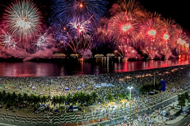 De colorés feux d'artifice éclatent dans le ciel nocturne au-dessus d'une baie parsemée de navires, observés par une foule immense sur une plage de sable.