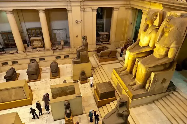 Una veduta dall'alto di una sala museale ricolma di colossali statue egizie antiche, sarcofagi e manufatti, con visitatori in basso.