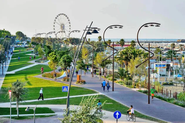 Le persone passeggiano e vanno in bicicletta lungo i sentieri di un parco costiero paesaggistico, illuminato da lampioni moderni e con una ruota panoramica in lontananza.
