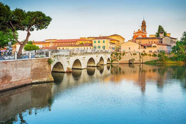 Un ponte in pietra a più arcate attraversa un fiume calmo, con colorati edifici storici e un campanile di chiesa che si riflettono nell'acqua.