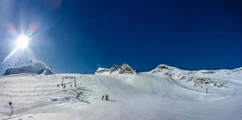 Un ampio pendio montano innevato con persone che sciano sotto un sole splendente e un cielo azzurro chiaro.