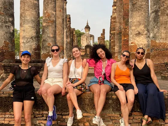 Eine WeRoad-Gruppe von sechs Frauen sitzt auf einer Ziegelmauer und posiert für ein Foto vor antiken Tempelruinen mit einer Buddha-Statue.
